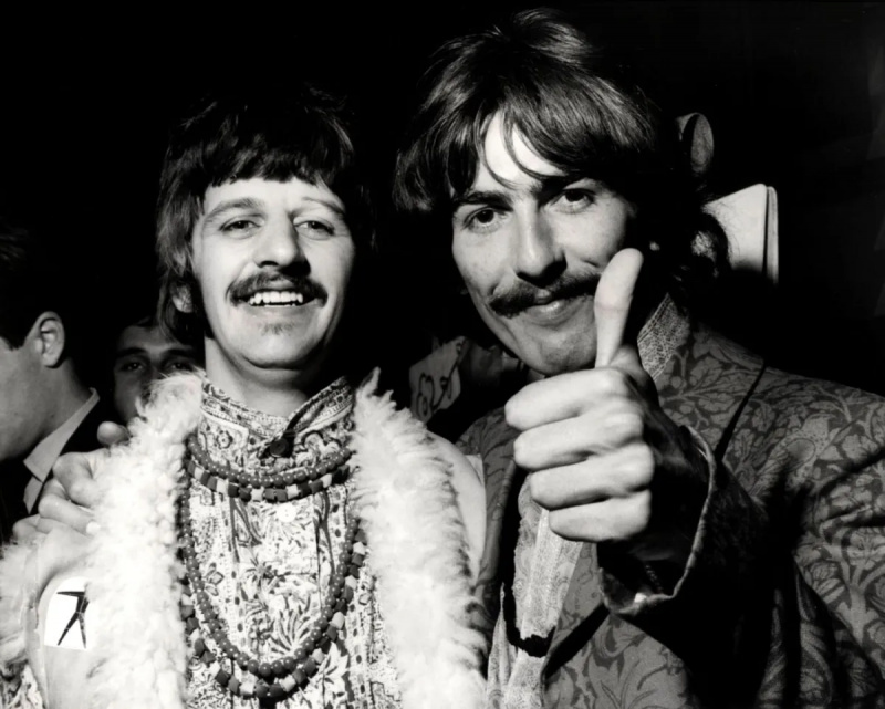   O imagine alb-negru cu George Harrison ridicând degetul mare cu brațul în jurul lui Ringo Starr's shoulders.