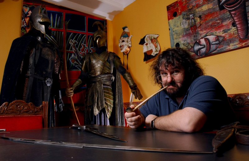   Peter Jackson poseert met'The Lord of the Rings' props with a pipe in his mouth. He's wearing a blue shirt and glasses with his arms resting on the table.